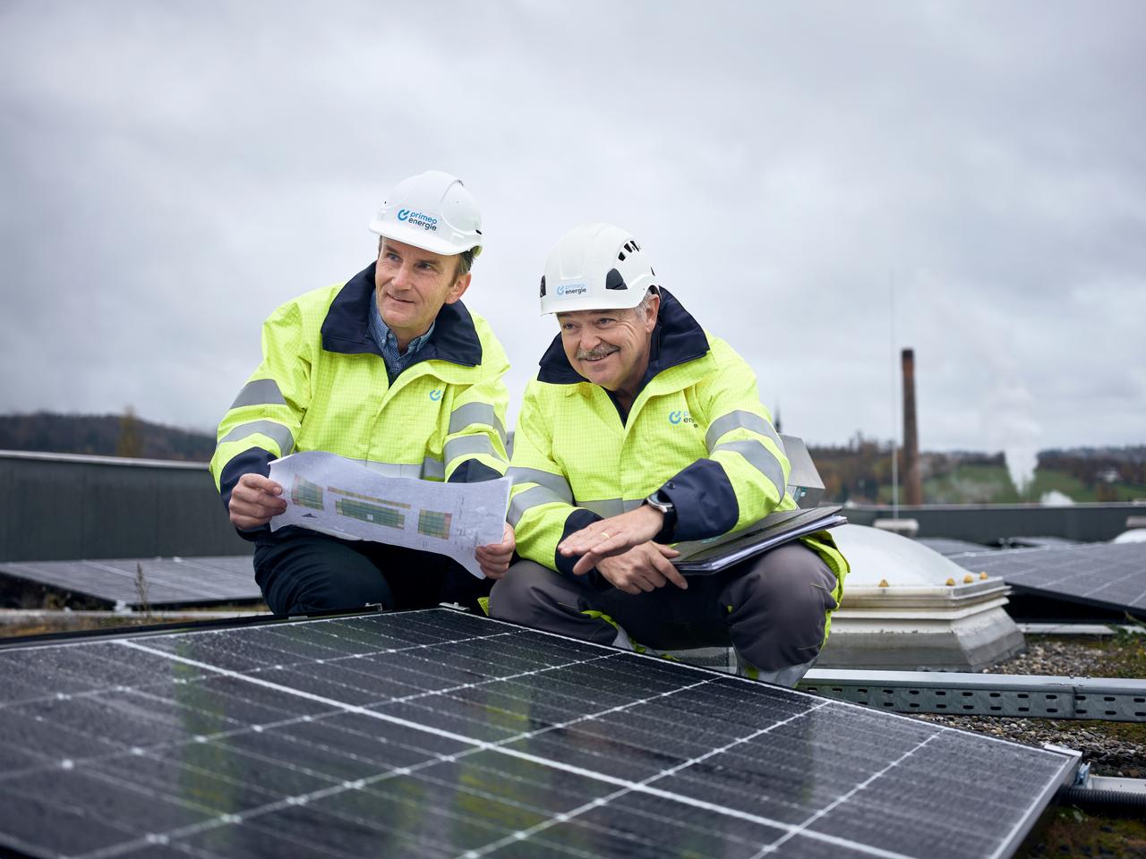  Zwei Primeo Energie Ingenieure in Warnwesten und Helmen überprüfen eine Solaranlage auf einem Dach. Sie studieren Baupläne und notieren Details. 