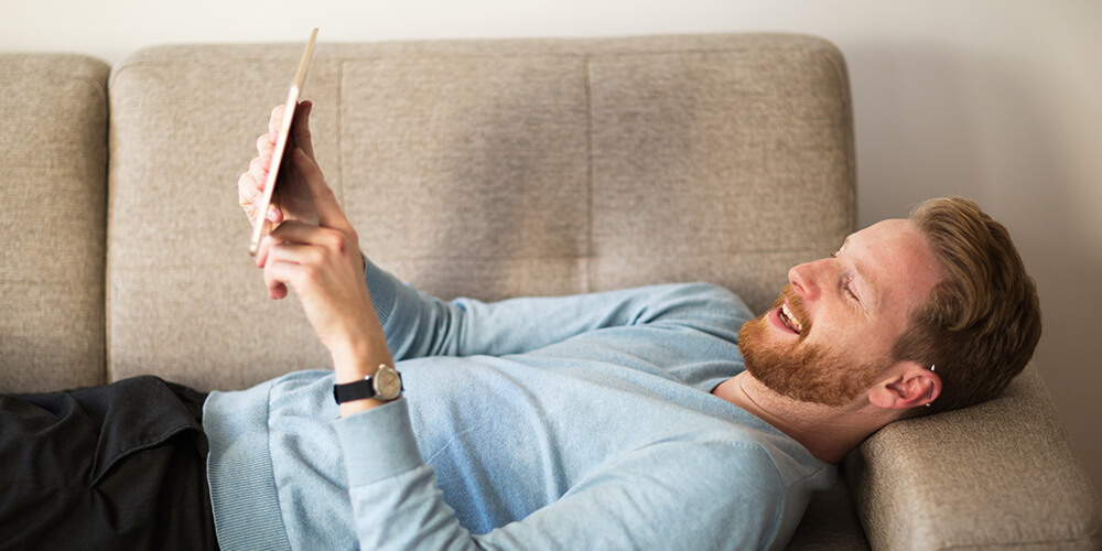 Ein lächelnder Mann liegt entspannt auf einem Sofa und hält ein Tablet in den Händen, während er darauf schaut. 